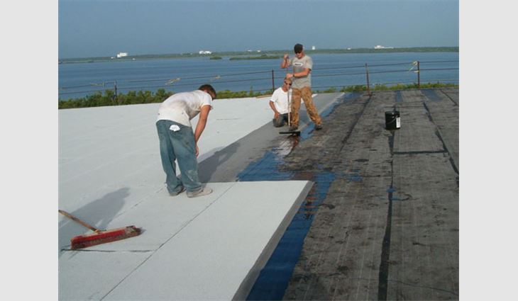 Roofing workers apply adhesive to the back of the cap sheet before installation.
