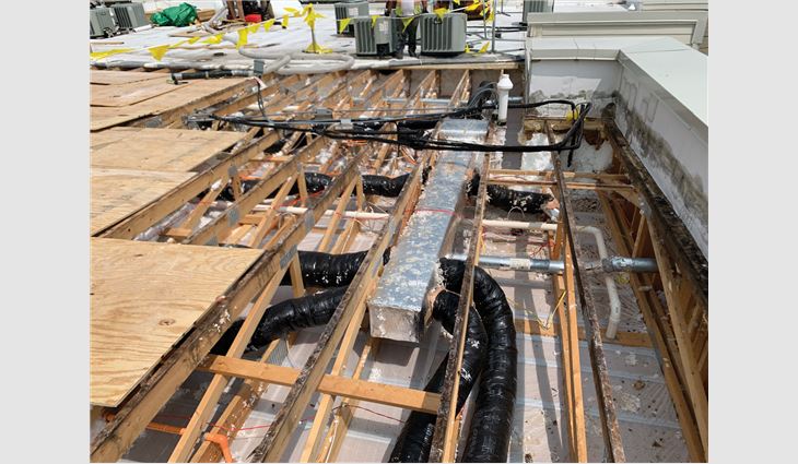 Photo 1: An overview of a cold roof being renovated. The blown insulation has been vacuumed out. The ductwork is visible running in the interstitial space. Although new plywood is being installed, the dark-colored top truss chord resulted from biological growth.